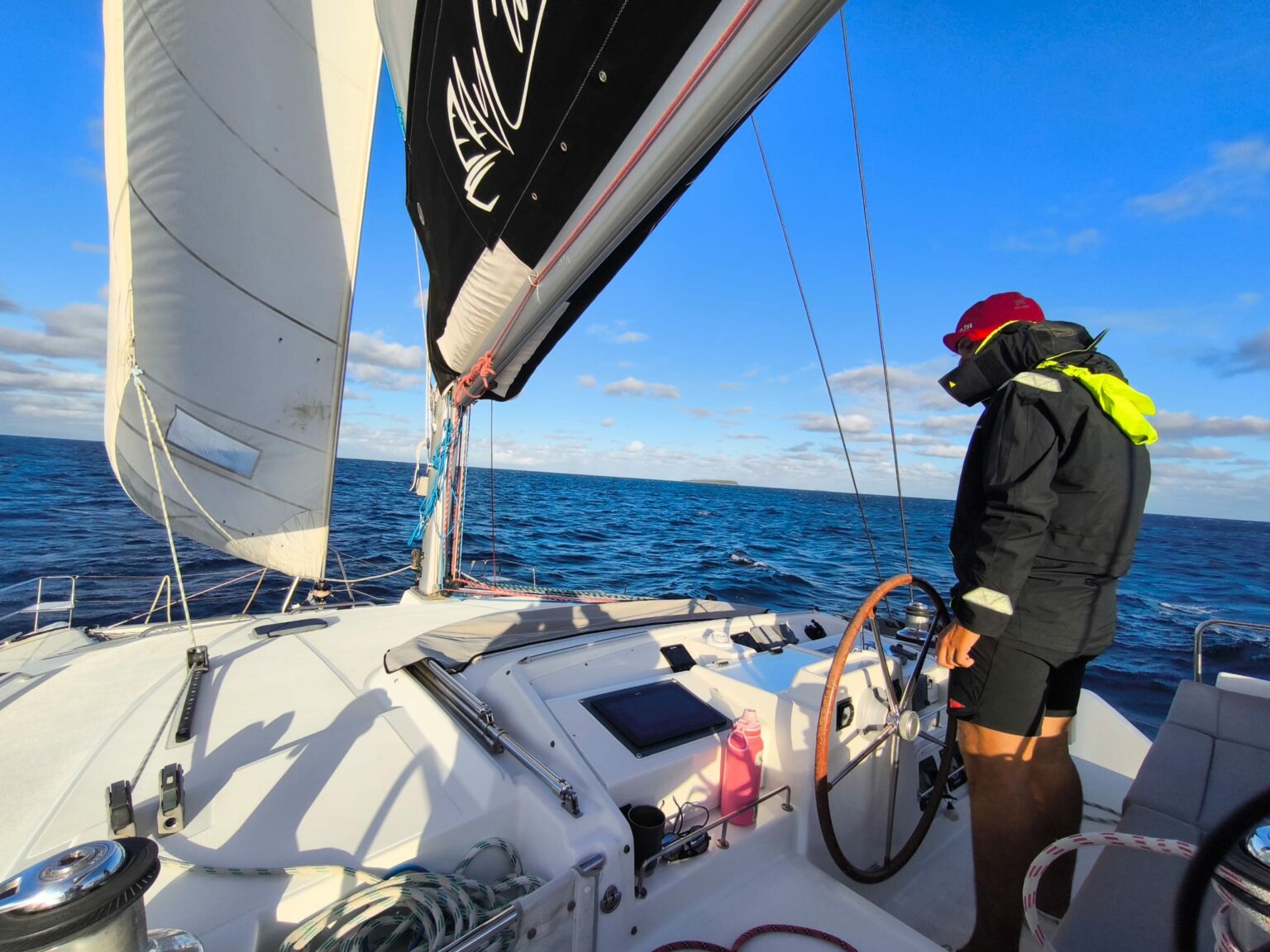 Un équipier en vêtement de quart à la barre d’un catamaran sous voiles, mer bleue et ciel avec de petits nuages