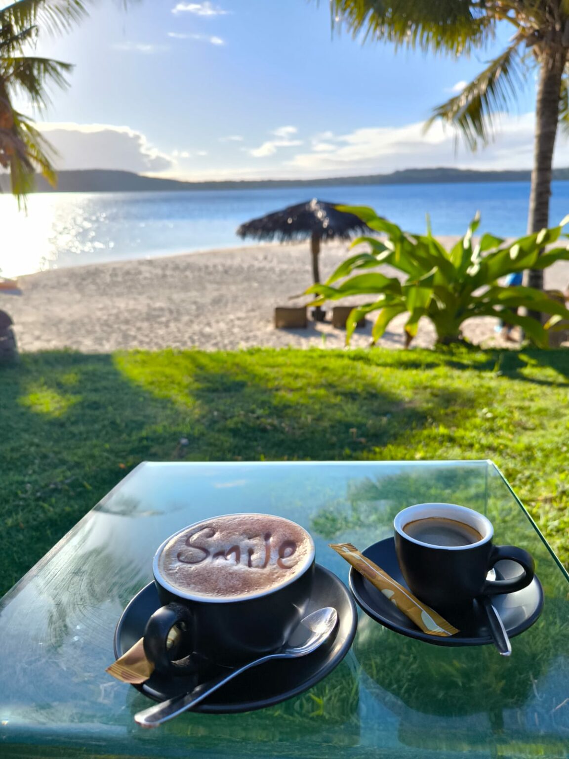 Two cups of coffee on a glass table, beach and palm trees behind