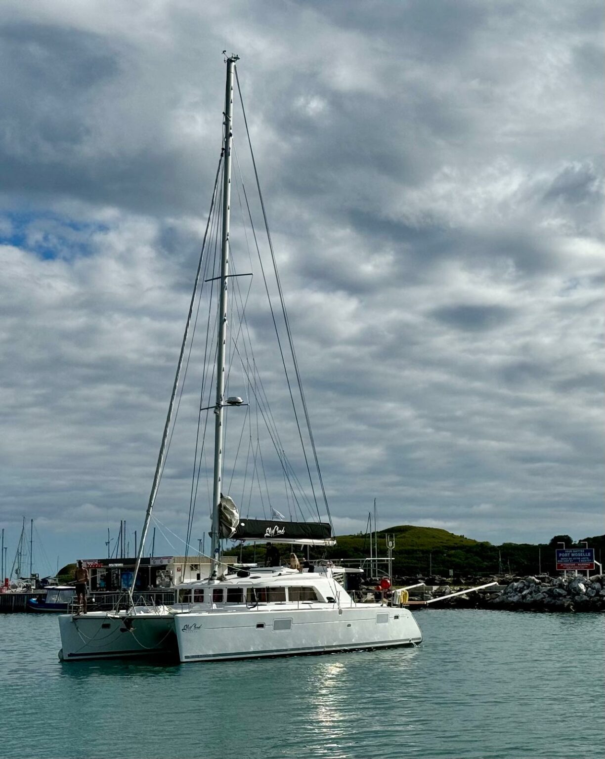 A moored sailing catamaran, tall mast, cloudy sky and hill behind