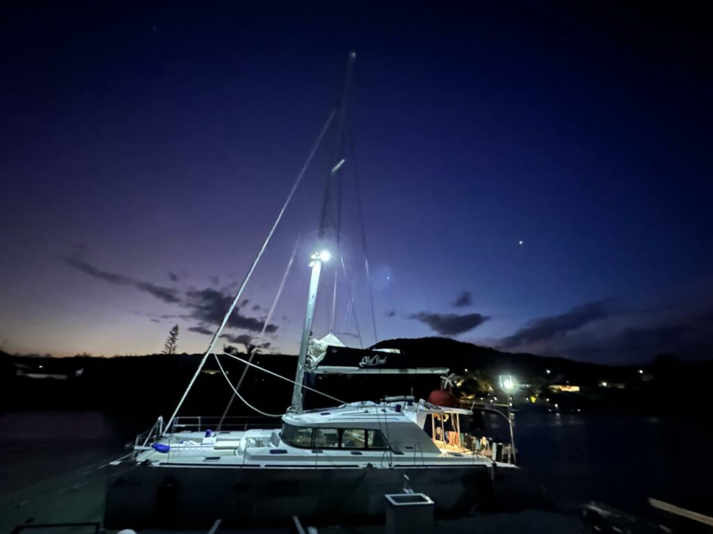 A catamaran moored at night, purple sky, mast lit by a lamp, dark shore behind