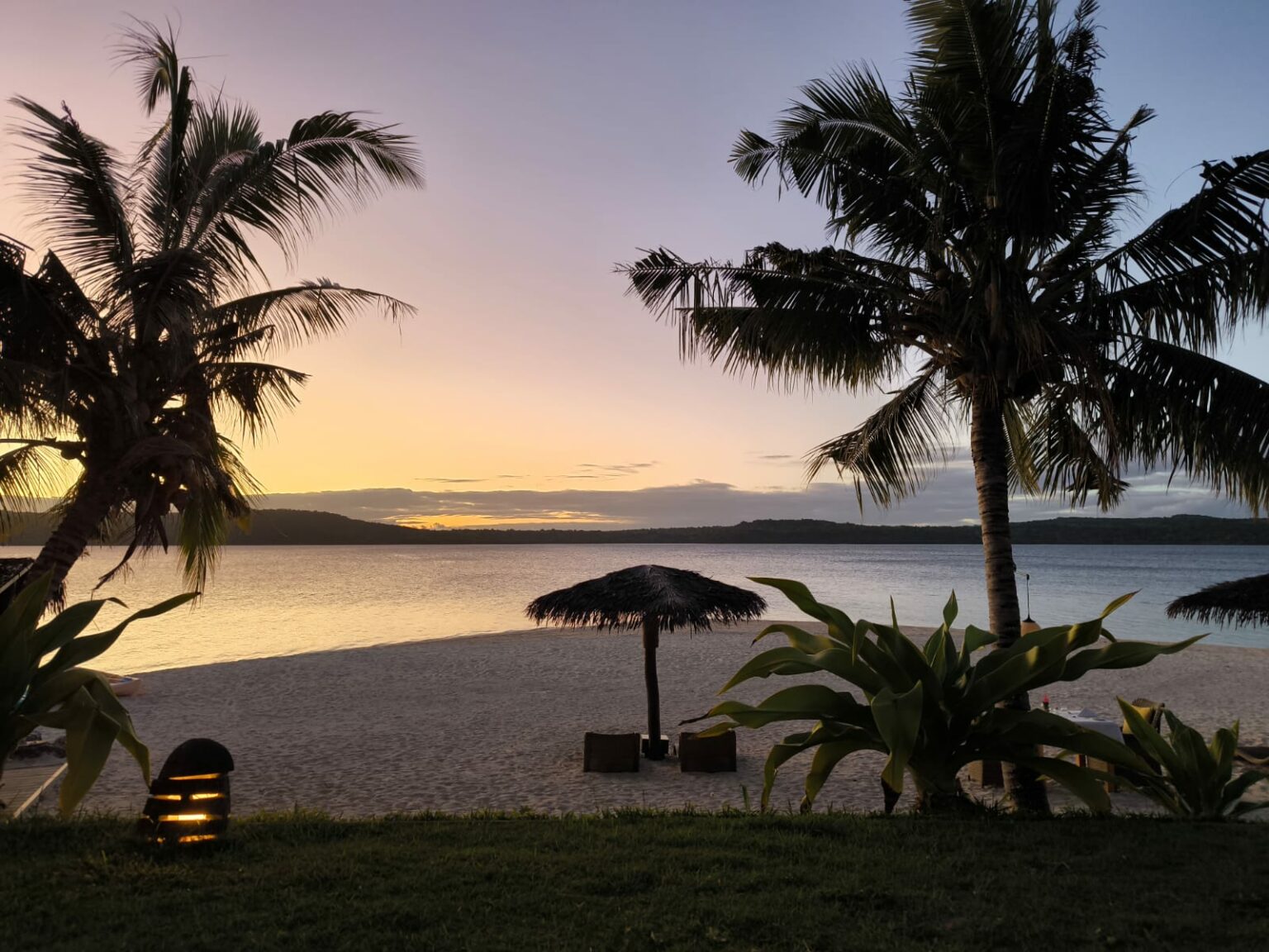 Silhouettes de cocotiers et parasol de paille devant une mer calme, ciel orangé au coucher du soleil