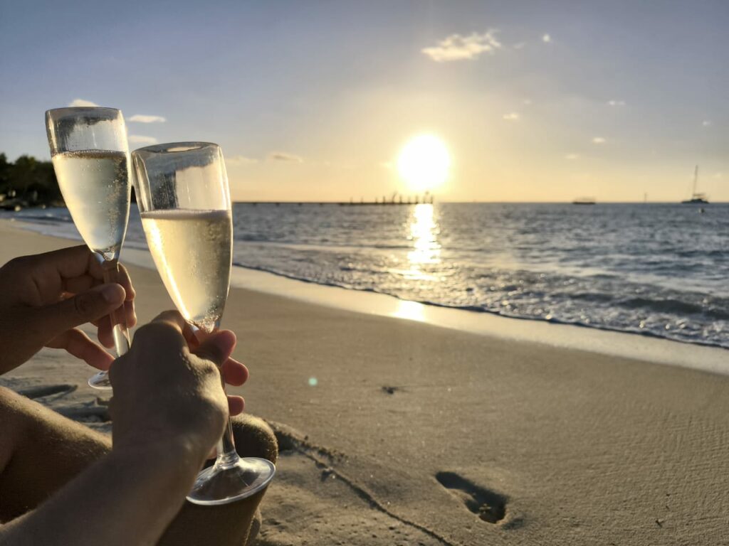 Deux coupes entrechoquées sur une plage, soleil bas et reflet sur l’eau, traces de pas dans le sable