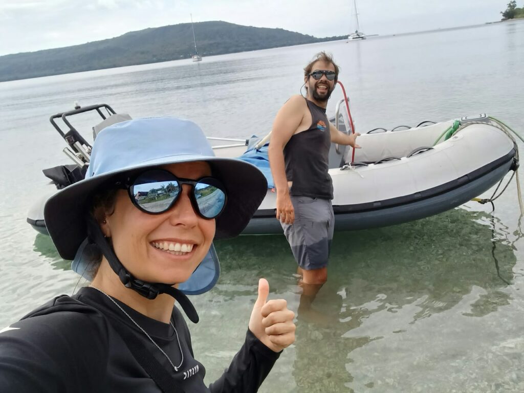 A smiling crew member in hat and mirrored glasses gives a thumbs up in front of a tender, another crew member standing in the water
