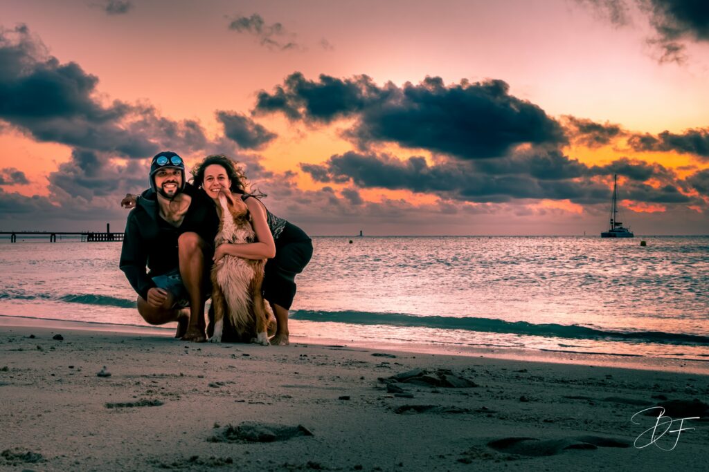 Deux équipiers et le chien sur une plage au coucher du soleil, un catamaran au mouillage derrière eux.