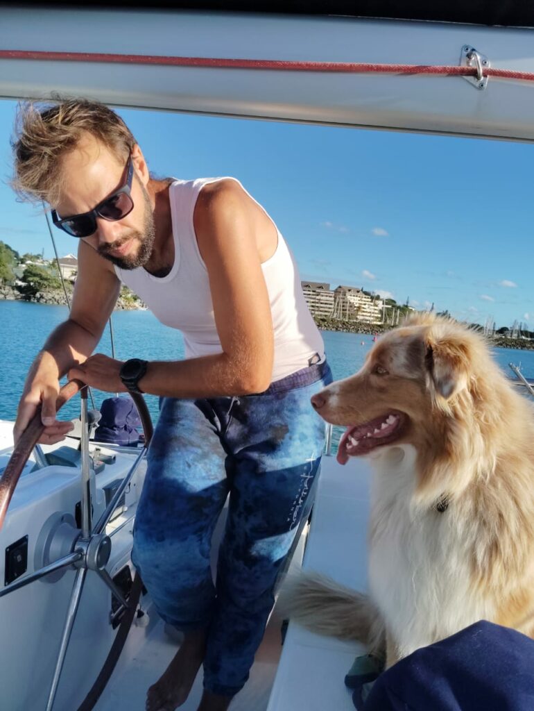 A crew member in sunglasses at the helm, the Australian shepherd sitting beside him, harbour behind