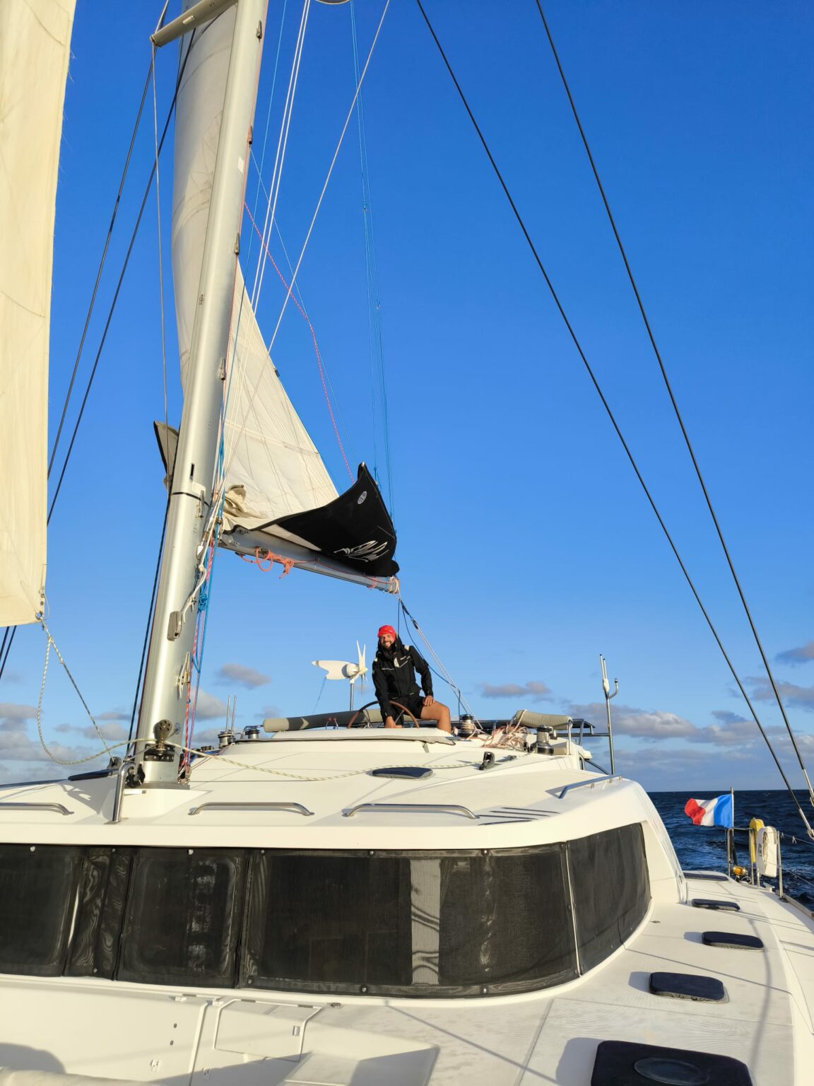 Un catamaran vu de l’avant, grand-voile hissée, un équipier sur le pont et le pavillon français à l’arrière