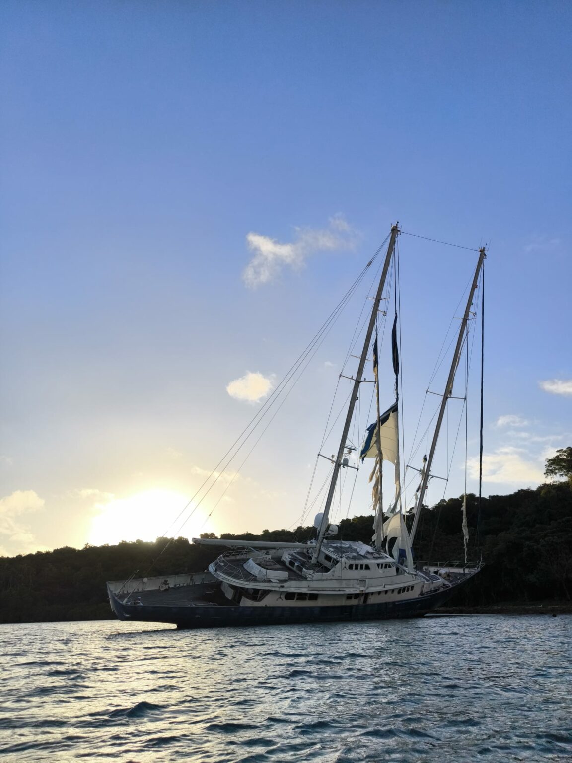 A ketch at anchor off a wooded shore, low sun on the left and calm water in the foreground