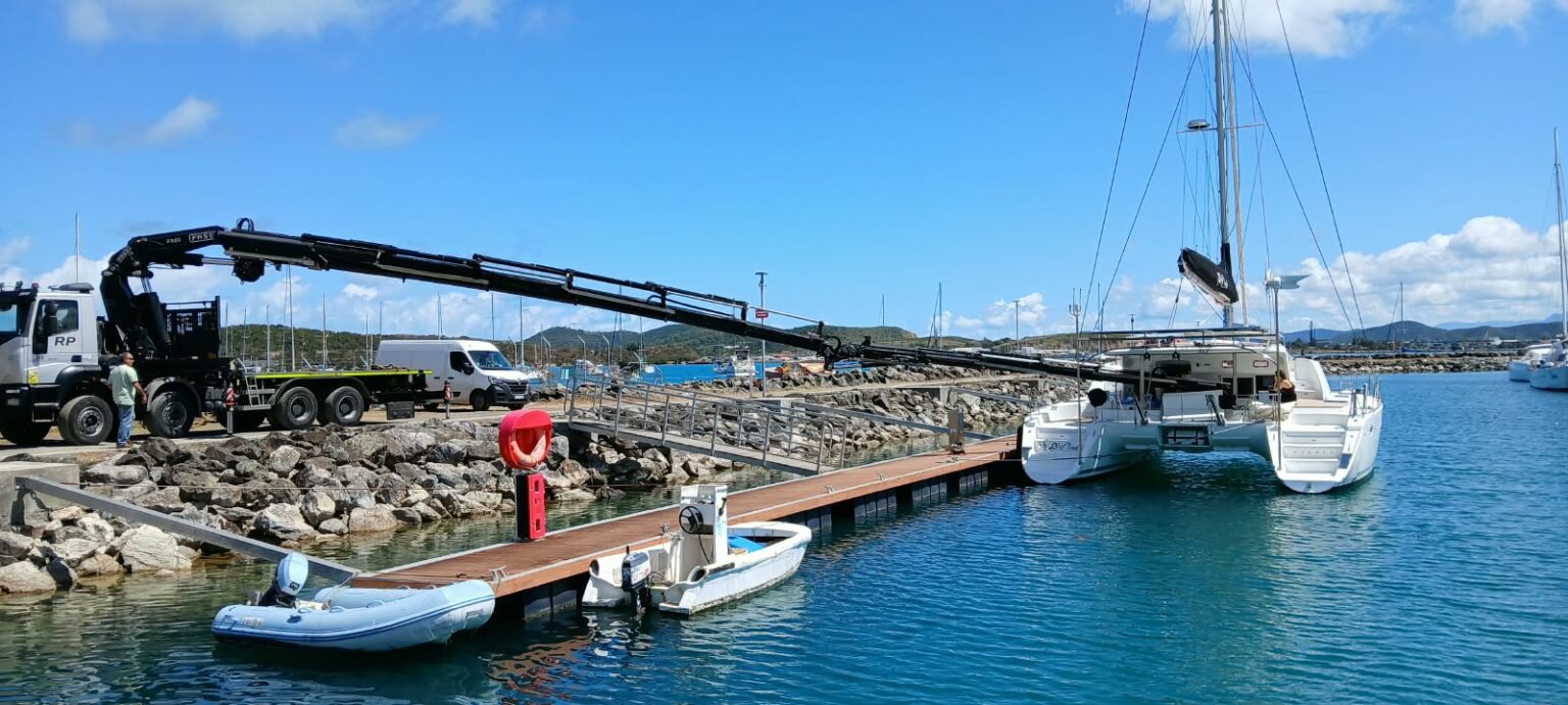 A harbour crane on a stone quay, a catamaran afloat and two tenders along the pontoon