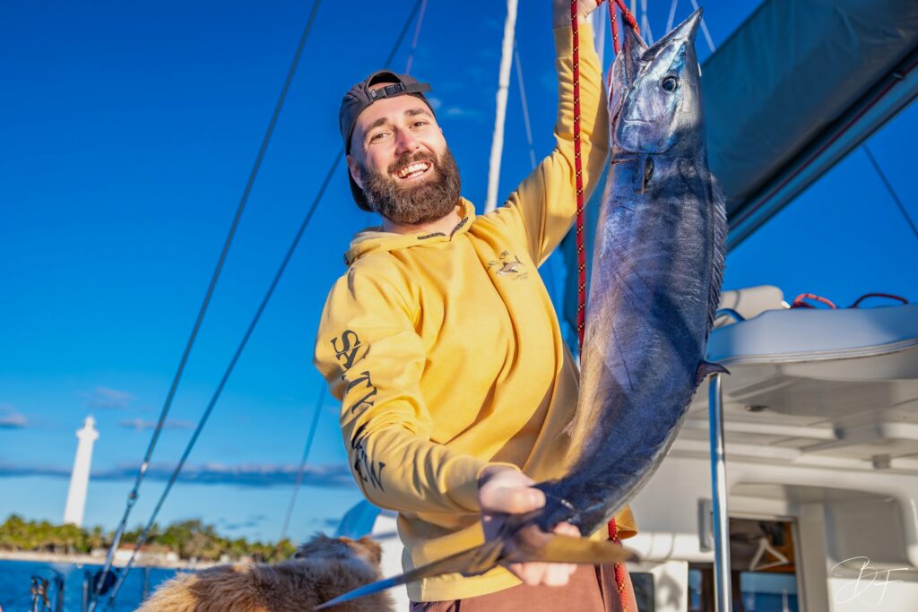 Un équipier présente une prise au retour de pêche, à bord de myCloud, phare en arrière-plan.