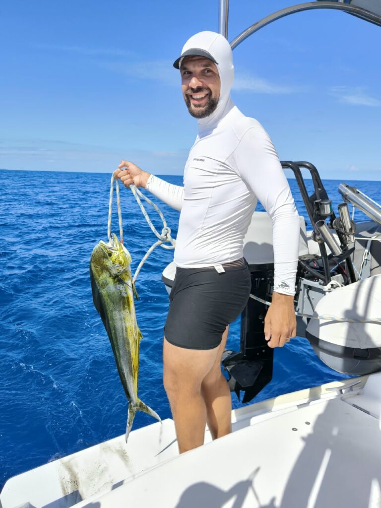 A crew member in a white rash guard holds a yellow mahi-mahi by the line, blue sea behind him