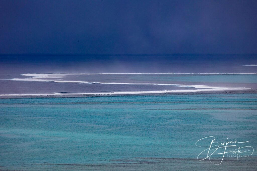 Ligne d’écume sur la barrière de corail, lagon turquoise au premier plan et grain sombre à l’horizon.