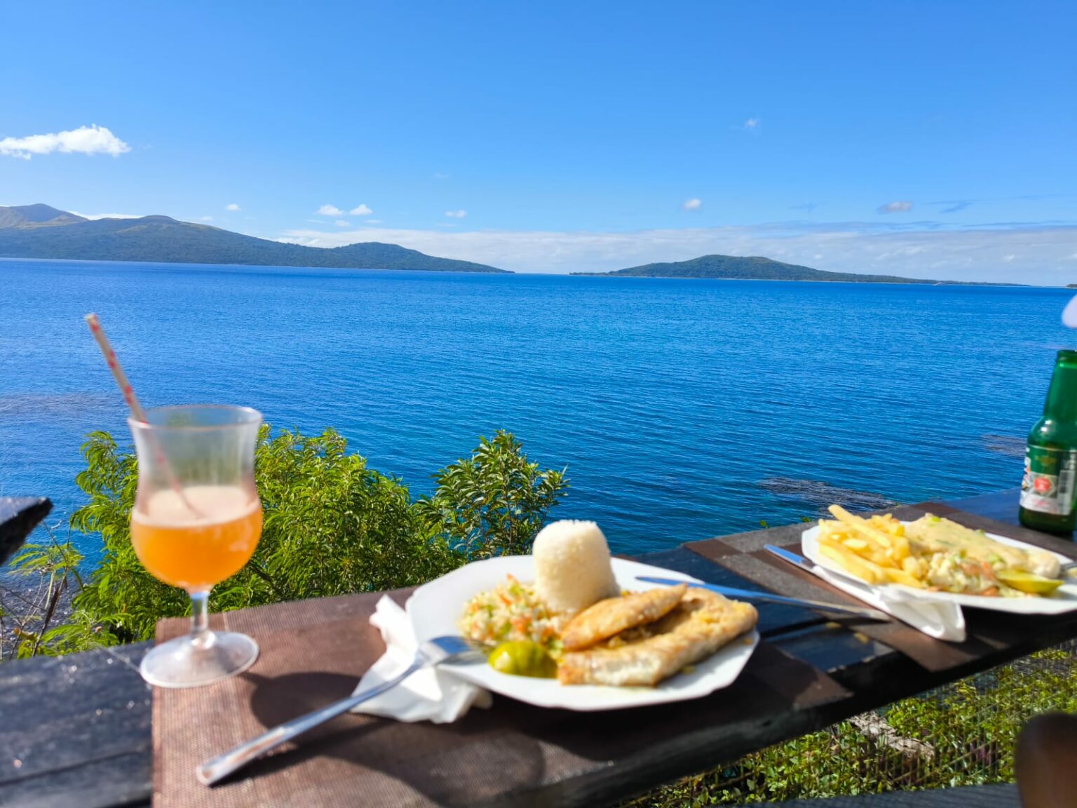 Une assiette de poisson frit, du riz et des frites avec un verre de jus d’orange, devant un lagon bleu