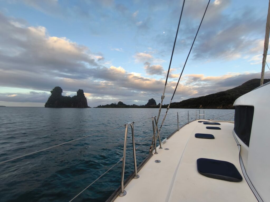 Le pont blanc d’un catamaran au premier plan, deux pitons rocheux et une côte sous un ciel nuageux