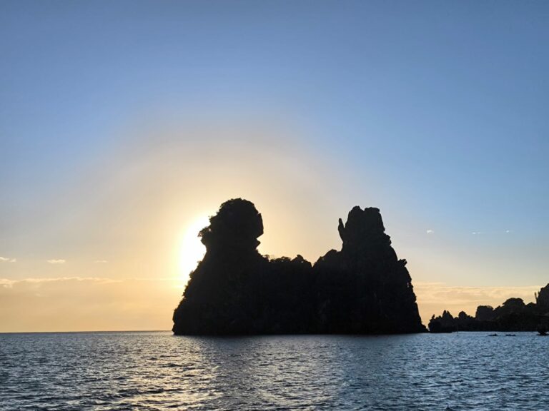 Silhouette sombre de deux rochers devant un soleil bas, halo doré sur une mer calme