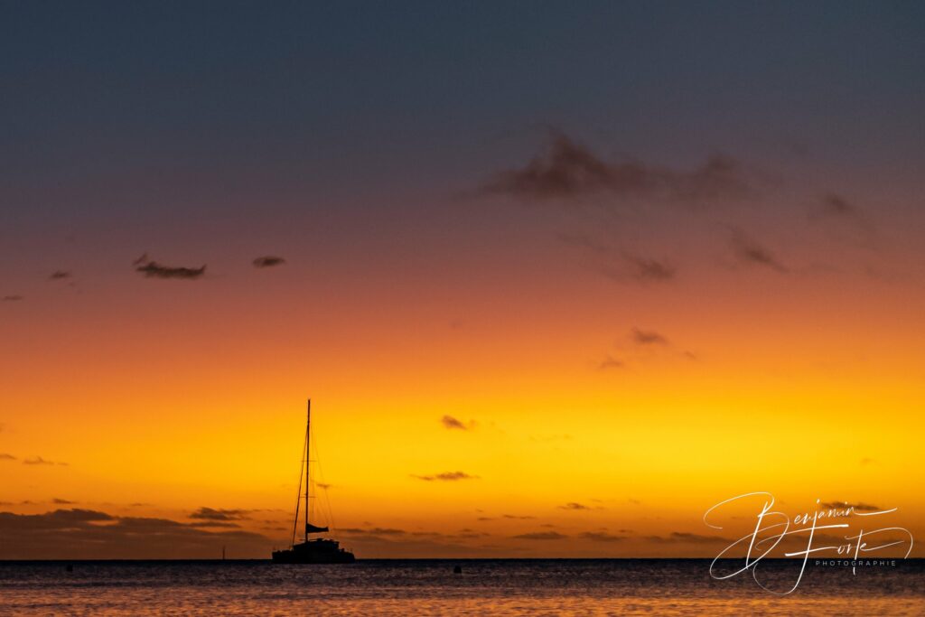 Silhouette d’un catamaran au mouillage devant un ciel orange, dégradé du bleu nuit à l’or.