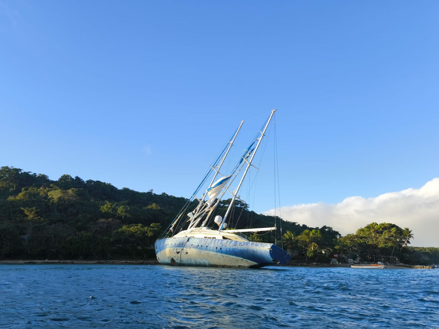 A sailboat with a weathered hull aground and tilted off a green shore, blue sea in the foreground